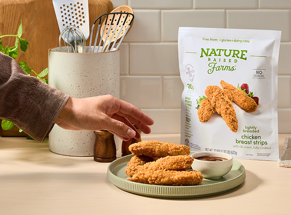 Hand reaching for breaded chicken strips on a plate, with Nature Raised Farms frozen chicken breast strips bag on a kitchen counter